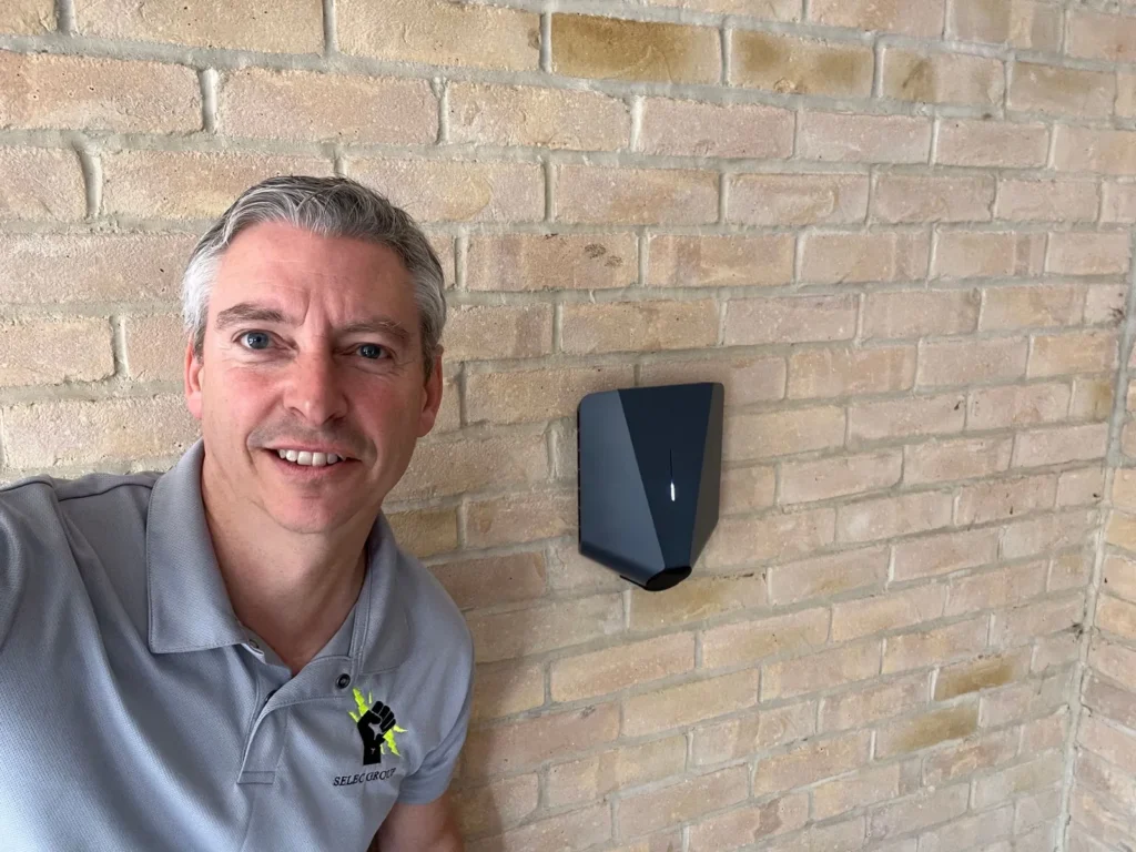 Jason Pope of Selec Group beside a black EV charger he fitted on a light brick house wall